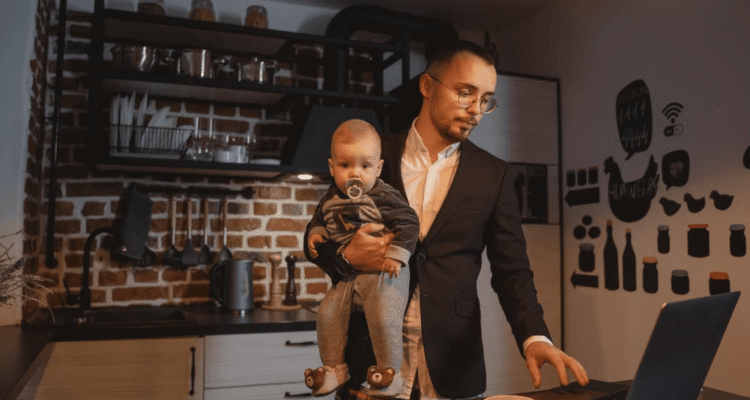 A parent entrepreneur uses a laptop while holding a baby
